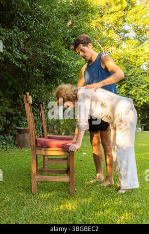 Physiothérapeute aide une femme âgée à effectuer des exercices de flexion à l'aide d'une chaise dans un jardin verdoyant luxuriant, favorisant la mobilité et le bien- Banque D'Images