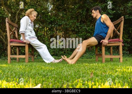 Femme âgée et son entraîneur personnel pratiquant des exercices de renforcement des jambes en utilisant des chaises dans un jardin, favorisant le vieillissement sain et la mobilité Banque D'Images