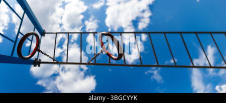 Vue rapprochée d'anneaux de gymnastique en métal et de barres d'échelle horizontales sur une aire de jeux, avec ciel bleu et nuages en arrière-plan. Banque D'Images