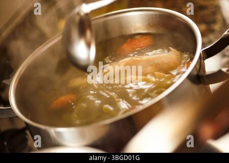 Chef ajoutant de l'eau dans une casserole en acier inoxydable avec le poulet et les légumes mijotant sur la cuisinière, créant un délicieux bouillon Banque D'Images