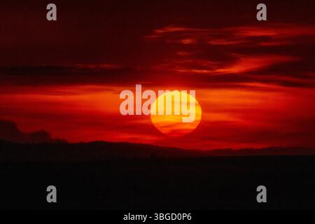 soleil d'été adiant brillant puissamment sur les collines silhouettes, encadré par un ciel sombre et de riches nuages rouge-orange lors d'un coucher de soleil saisissant en soirée Banque D'Images