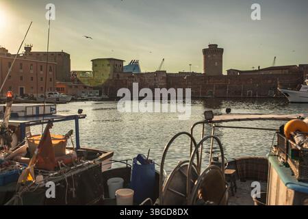 Livourne, Italie 1er janvier 2025 : bateaux de pêche amarrés dans le port de livourne, italie, pendant le coucher du soleil, avec la fortezza vecchia en arrière-plan Banque D'Images