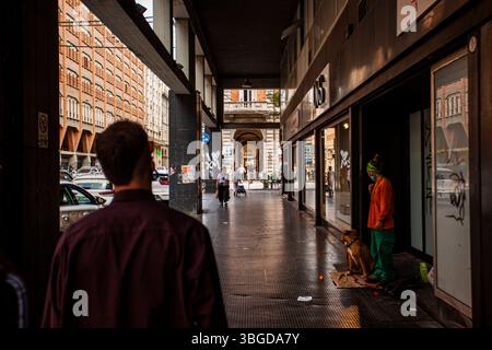 Bologne, Italie 1er janvier 2025 : homme marchant sur un trottoir couvert à bologne, italie, passant par des magasins et un sans-abri avec un chien Banque D'Images