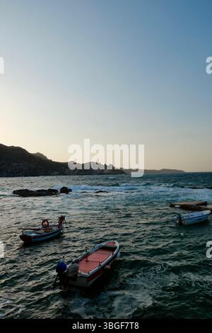 Plusieurs petits bateaux de pêche flottant dans un port calme pendant le coucher du soleil, avec de douces ondulations sur l'eau et des collines en arrière-plan, probablement sur un méditer Banque D'Images