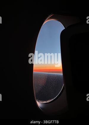 Vue de la fenêtre de l'avion au coucher du soleil, montrant des gradients orange et bleu vifs au-dessus de l'horizon en vol. Banque D'Images