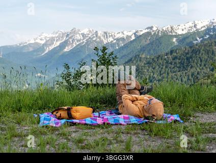 randonneur masculin en sweat à capuche jaune avec sac à dos reposant sur une couverture bleue sur l'herbe sur fond de chaîne de montagnes avec des sommets enneigés, randonnée et nat Banque D'Images
