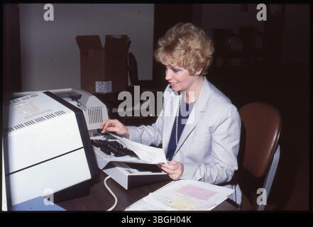 Une employée de bureau non identifiée utilise un clavier d'ordinateur dans un environnement de bureau entre 1981 et 1990. Une imprimante avec du papier en continu est visible en arrière-plan. Banque D'Images