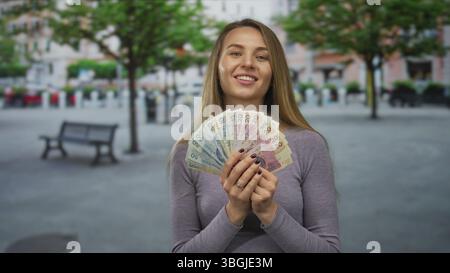 Femme souriante et tenant des billets de banque en zloty polonais dans une rue de la ville, mettant en valeur la richesse dans un fond urbain en plein air. Banque D'Images