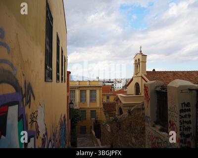 Rue calme dans le quartier de Plaka, Athènes, avec des murs jaunes recouverts de graffitis, une petite église et des paysages authentiques sous un ciel nuageux. Banque D'Images