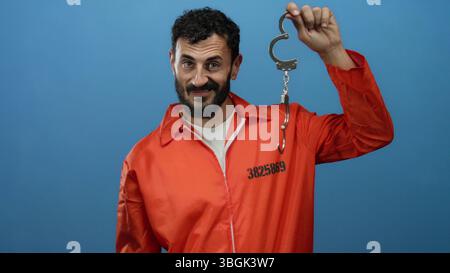 Homme hispanique avec la barbe dans la combinaison de prison orange tenant les menottes sur fond bleu, représentant le thème de l'incarcération. Banque D'Images
