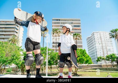 Deux patineurs joyeux avec des casques et des équipements de protection tenant des planches à roulettes se sourient l'un à l'autre dans un skate Park urbain Banque D'Images
