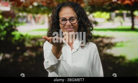 Femme avec des cheveux bouclés et des lunettes étend la main vers la caméra dans un cadre de parc extérieur vibrant, exsudant la chaleur et la convivialité. Banque D'Images