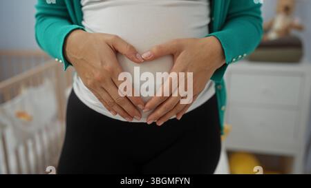 Femme enceinte dans la chambre berce doucement le ventre avec des mains aimantes formant le symbole du cœur dans l'intérieur chaleureux et chaleureux près d'un berceau de bébé confortable. Banque D'Images