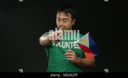 Jeune homme en chemise verte tenant le drapeau philippin sur fond noir faisant des gestes de la main, représentant le bénévolat et la fierté nationale. Banque D'Images