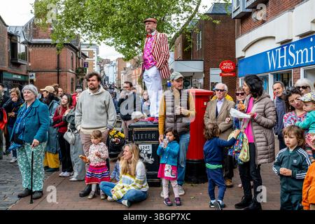 Un homme habillé de couleurs vives se tient sur Une poubelle à litière pour Une meilleure vue d'Un groupe d'artistes de rue du jour de mai, High Street, Lewes, East Sussex, Royaume-Uni. Banque D'Images