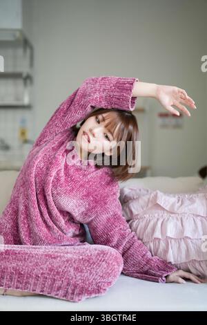 Dans une pièce calme et bien éclairée, une femme japonaise enceinte au début de la vingtaine aux cheveux bruns courts, portant une robe en tricot rose, est assise sur un canapé pratiquant Banque D'Images