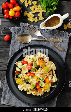Salade de pâtes farfalle méditerranéenne avec tomates cerises, courgettes, poivrons et olives dans un bol noir, avec huile et vinaigrette aux herbes. Lumière naturelle, réelle Banque D'Images