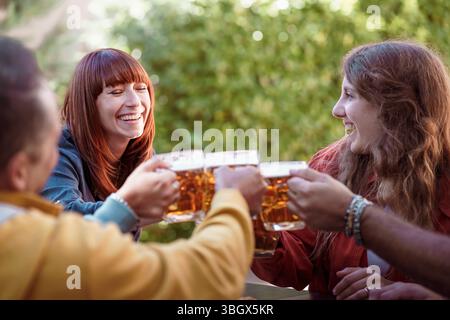 Un groupe de jeunes amis heureux rit et prend des verres, toast avec des tasses de bière fraîche dans un jardin de pub extérieur ou un festival d'été. Momen authentique Banque D'Images