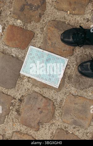 Une vue de haut en bas montre un puzzle de mots croisés partiellement terminé reposant sur un pavé texturé, avec les pointes des chaussures sombres d'une personne visible Banque D'Images