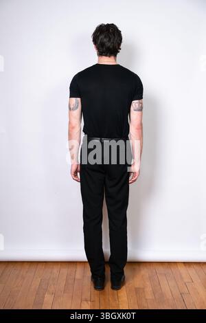 Portrait en studio d'un homme vu de derrière, vêtu d'un t-shirt noir et d'un pantalon sur fond blanc, montrant des tatouages sur les deux bras Banque D'Images