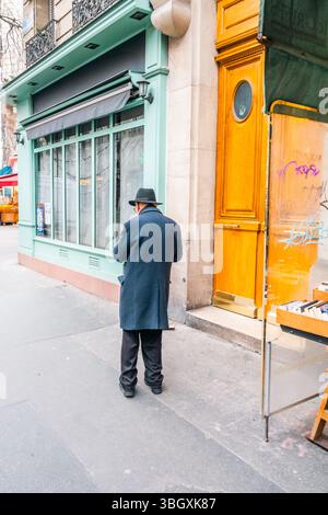 Un homme portant un pardessus sombre et un chapeau de style Fedora se tient sur un trottoir de la ville, vu de derrière. Il semble regarder vers un esprit de magasinage Banque D'Images