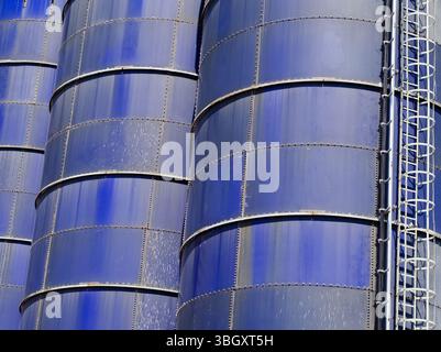 Un gros plan détaillé de plusieurs silos industriels bleus, mettant en évidence leurs panneaux métalliques rivetés et une échelle d'accès industrielle, sous un soleil fort Banque D'Images