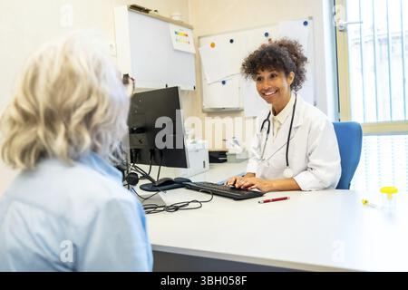 Médecin féminin souriant portant une blouse de laboratoire et tapant stéthoscope sur le clavier de l'ordinateur tout en écoutant la patiente senior dans la chambre d'hôpital Banque D'Images
