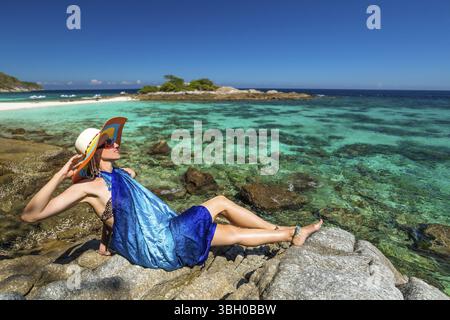 Vacances d'été dans la mer d'Andaman. Femme attrayante avec sarong bleu assis sur les rochers regardant la mer turquoise tropicale. Île de Racha Noi, Rawai, Tha Banque D'Images
