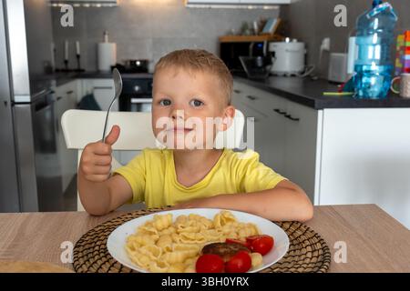 Garçon souriant en t-shirt jaune assis à la table de la cuisine avec assiette de pâtes et de légumes, tenant la cuillère et montrant les pouces vers le haut. Concept de sain Banque D'Images