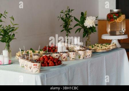 Une table à desserts magnifiquement agencée présente des parfaits de baies superposés, des pâtisseries variées, des fraises fraîches et un distributeur d'eau avec des agrumes, Banque D'Images