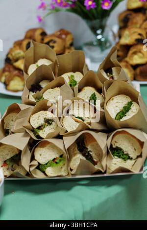 Une vue rapprochée montre des mini-sandwichs soigneusement disposés enveloppés dans du papier artisanal, accompagnés de pâtisseries appétissantes sur une nappe verte douce. flore délicate Banque D'Images