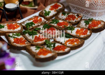 Un gros plan présente des rangées de canapés de pain de seigle foncé recouverts de fromage blanc à la crème, de caviar rouge vif et de persil frais. un hors-d'œuvre luxueux Banque D'Images