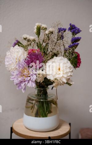 Un bouquet de fleurs sauvages joliment arrangé avec des dahlias violets et blancs et d'autres fleurs se tient dans un vase en verre transparent sur une petite table en bois, capturin Banque D'Images