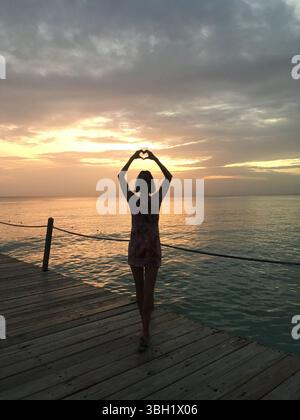 Silhouette de femme mince debout devant la vue spectaculaire sur la mer du coucher du soleil en République dominicaine, tout en faisant une forme entendue avec ses mains au-dessus de la tête Banque D'Images