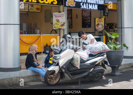 Jakarta, Indonésie - 9 février 2025 : homme debout à côté de la moto avec un sac à l'arrière. Une femme et un enfant sont assis sur le trottoir à côté du Banque D'Images