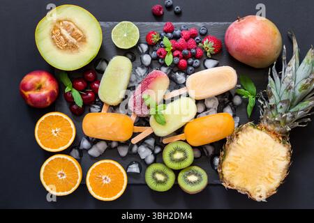 Au centre sur des glaçons une variété de cônes de sorbet de fruits autour des ingrédients fruits et baies sur un fond noir. Variété de cônes de sorbet de fruits et baies. Horizontal. Vue de dessus. Banque D'Images
