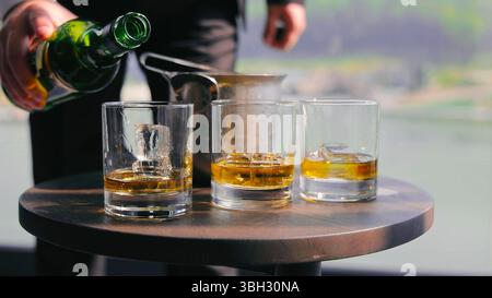 Verser le whisky dans des verres avec de la glace sur la table en bois. Un gros plan de whisky versé dans trois verres transparents avec des glaçons sur une table en bois. Banque D'Images