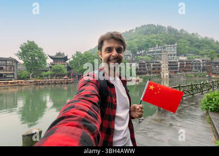 Profiter de vacances en Chine. Voyages et technologie. Jeune homme avec le drapeau chinois national prenant selfie à Fenghuang Banque D'Images