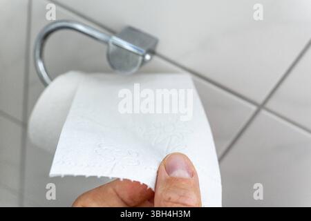 Bavière, Allemagne - 24 mai 2025 : une main tend pour le papier toilette sur le support dans la salle de bain - un moment quotidien dans le ménage qui représente visuellement le sujet de l'hygiène, de la fonctionnalité et des exigences sanitaires. *** Eine hand greift nach Toilettenpapier an der Halterung im Badezimmer ein alltäglicher moment im Haushalt, der das Thema Hygiene, Funktionalität und Sanitärbedarf visuell darstellt. Banque D'Images