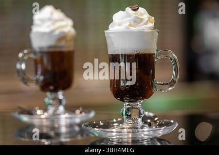 Bavière, Allemagne - 05 mai 2025 : décoré d'une garniture de crème savamment pulvérisée et de grains de café, le café dans un verre se présente comme une boisson chaude élégamment servie pour des moments de détente dans un cadre moderne. *** Mit einer kunstvoll aufgespritzten Sahnehaube und Kaffeebohnen verziert, präsentiert sich der Kaffee im Glas als stilvoll serviertes Heißgetränk für entspannte Momente in modérner Umgebung. Banque D'Images