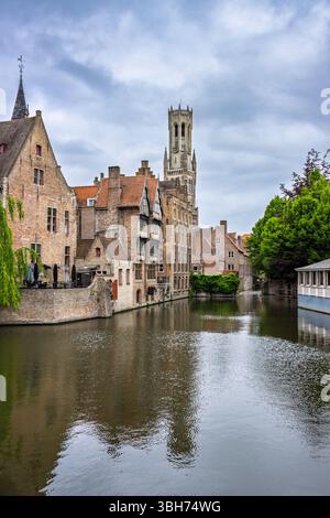 Le célèbre beffroi médiéval de Bruges s'élève au-dessus des eaux pittoresques du canal, avec des bâtiments traditionnels en briques et des arbres au premier plan. Banque D'Images