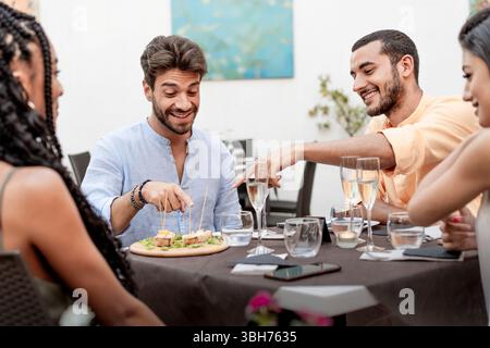 Un groupe diversifié de jeunes amis heureux partagent des hors-d'œuvre et du vin mousseux, socialisant dans un élégant restaurant ou bar. Une joyeuse scène d'amitié Banque D'Images