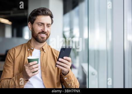 Homme d'affaires masculin réussi à l'intérieur du bureau debout par la fenêtre avec le téléphone dans les mains et tasse de boisson chaude. Homme d'affaires naviguant sur les médias sociaux et tapant un message texte. Banque D'Images