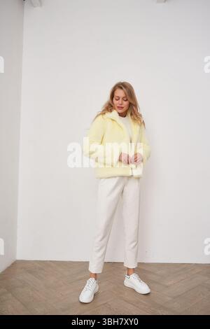 Femme blonde debout dans un coin de la pièce lumineuse portant une veste jaune confortable, un pantalon blanc et des baskets, regardant vers le bas avec une expression détendue. Lumière Banque D'Images