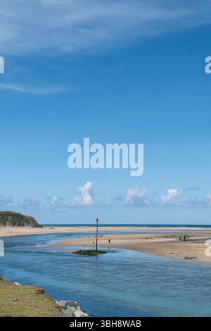 Un canal de marée calme coule à travers l'estuaire sablonneux à Hayle, Cornwall, sous un ciel bleu clair avec des personnages lointains marchant le long du rivage. Banque D'Images