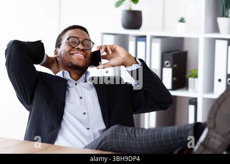 Manager noir décontracté assis avec les jambes sur le bureau, ayant la conversation Banque D'Images
