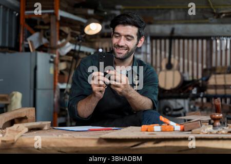 Joyeux charpentier assis dans un atelier de menuiserie à l'aide d'un smartphone. Concept de propriétaire de petite entreprise gérant le marketing, les médias sociaux, la communication, et Banque D'Images