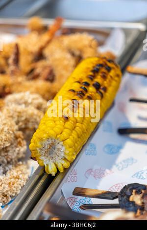 Gros plan d'un maïs grillé juteux sur l'épi, une collation populaire de Street food, avec des crevettes frites en arrière-plan. Assortiment de Street food. Prise de vue verticale wi Banque D'Images