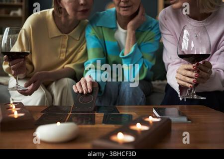 Trois femmes caucasiennes d'âge moyen assises ensemble tenant des verres de vin rouge, se livrant à la lecture de cartes de tarot à table avec des bougies, se concentrant sur les cartes et la conversation Banque D'Images