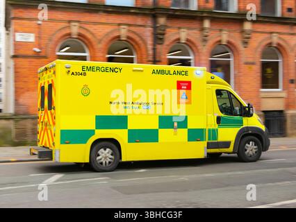 Une ambulance d'urgence jaune du service d'ambulance du Nord-Ouest conduisant à grande vitesse à travers une rue de la ville avec des bâtiments en briques. Banque D'Images